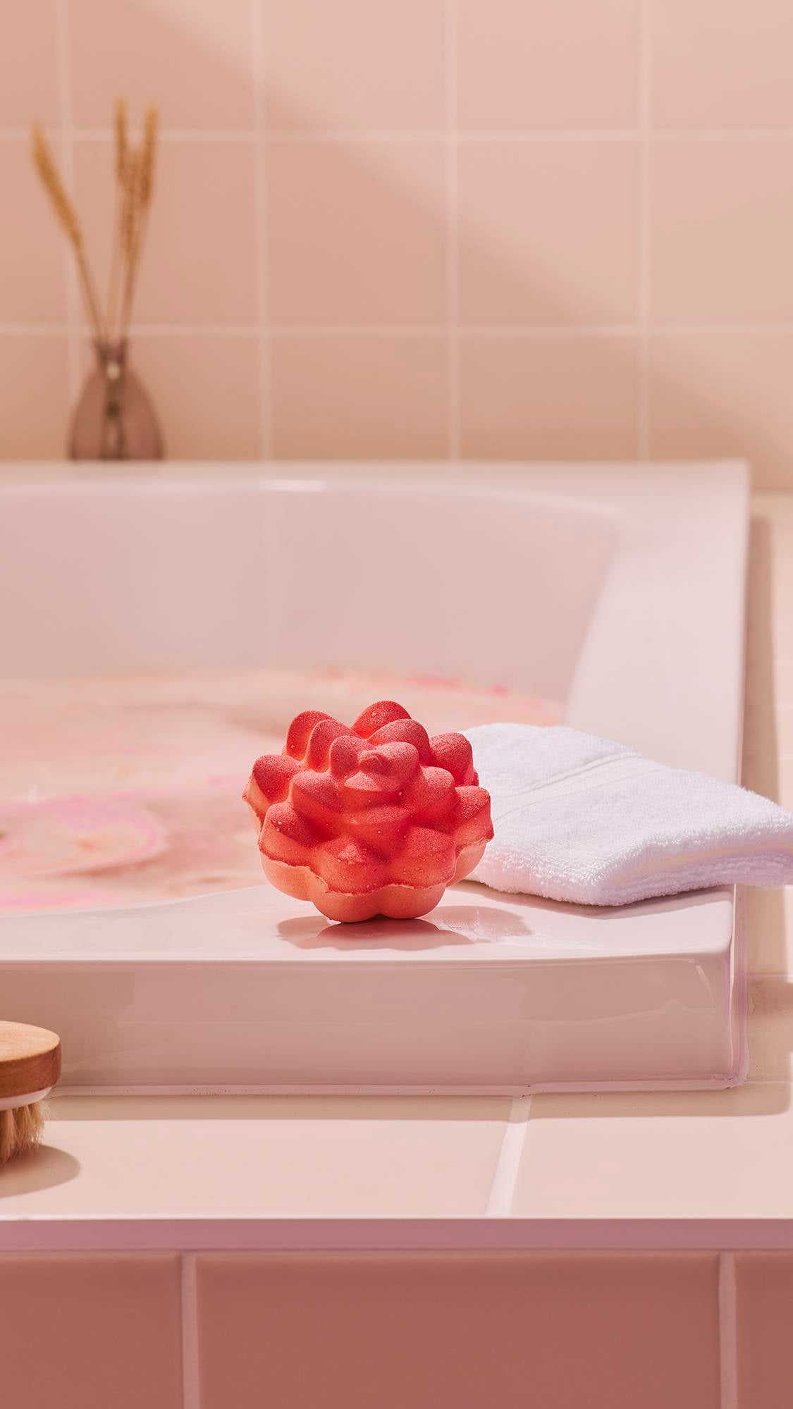 A vibrant coral-coloured "Comforter Rose" bath bomb sits on the edge of a white bathtub. In the background, a white flannel and a small vase with dried wheat create a relaxing spa-like atmosphere against pale beige wall tiles.