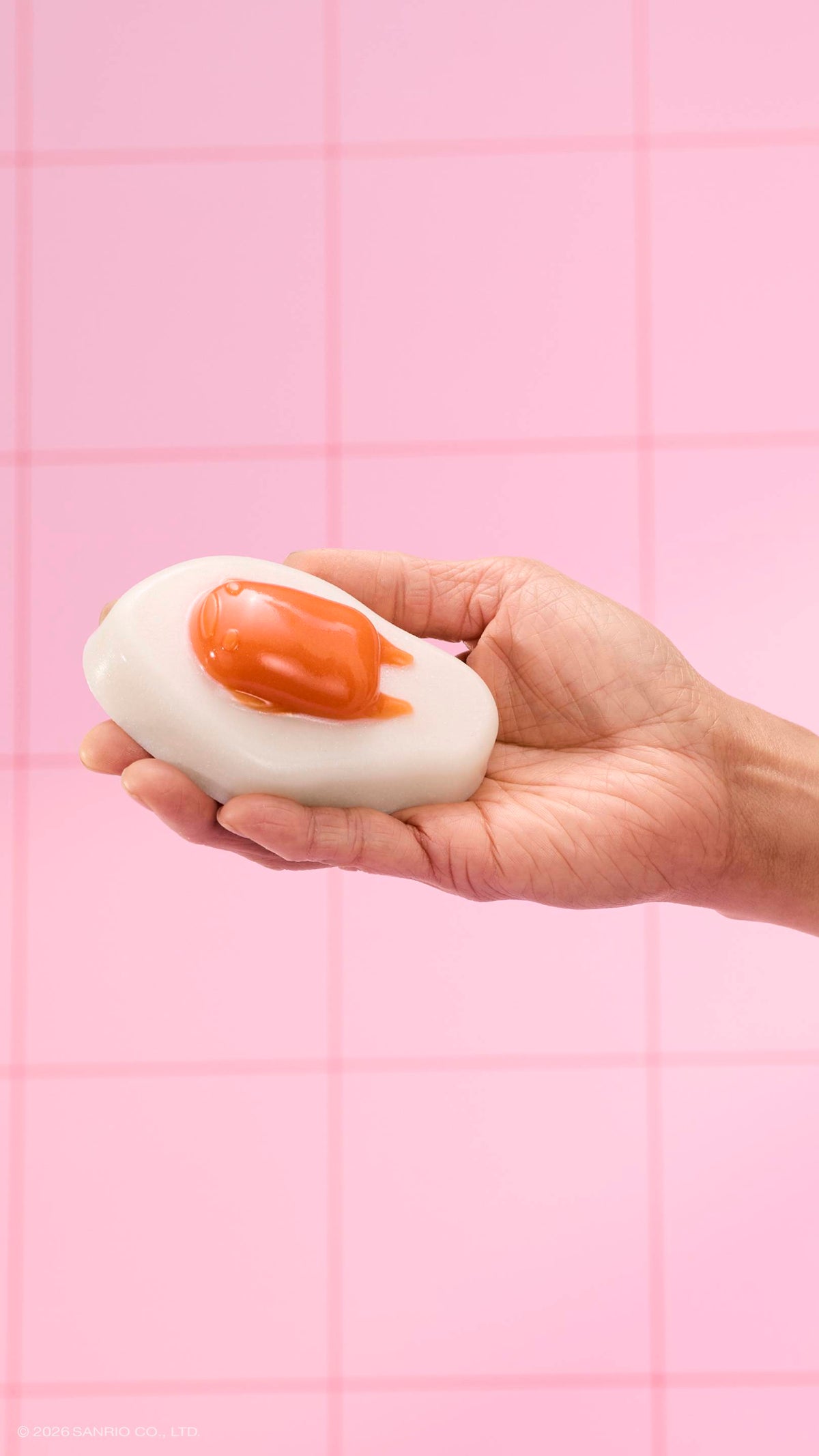 A hand holds a glossy, fried-egg-shaped shower jelly against a background of pink square tiles. The character Gudetama is featured as the orange "yolk" on the white jelly base.