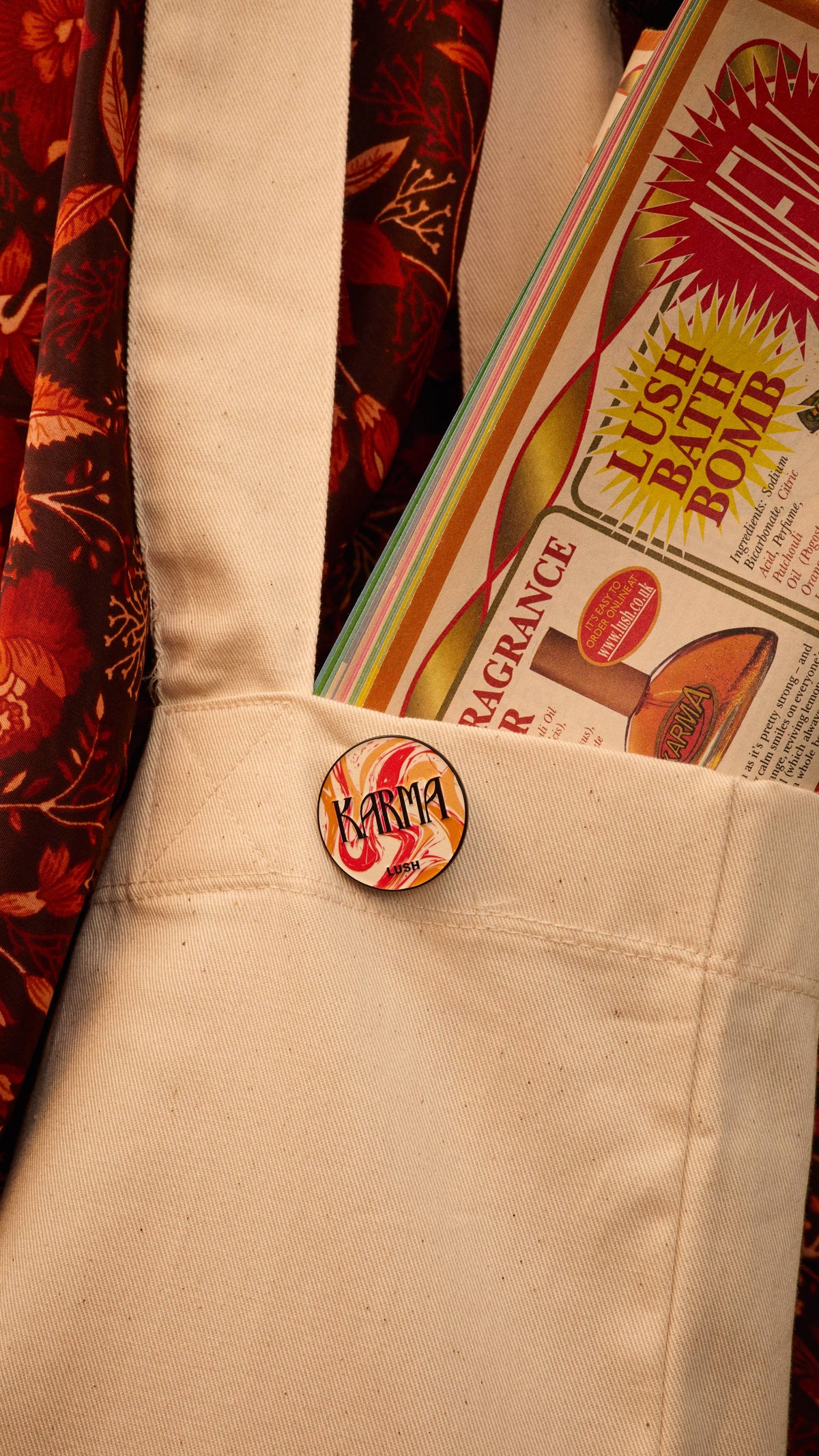 The image shows the red, orange and white marbled design Karma pin badge attached to a light canvas tote bag pocket. A colourful Lush magazine is visible inside, with a patterned fabric backdrop.