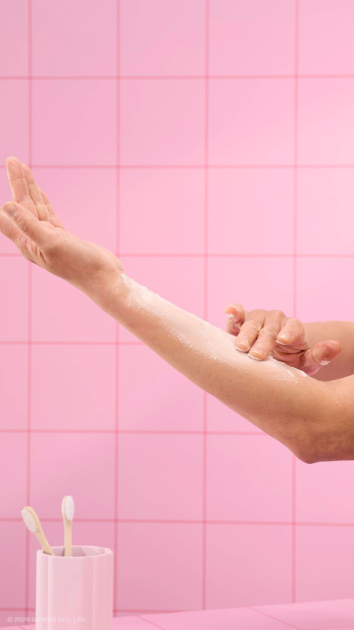 A close-up shows hands gently rubbing white dusting powder into an arm. The scene is set against a background of pink square tiles, with a pink toothbrush holder containing two toothbrushes in the lower-left corner.