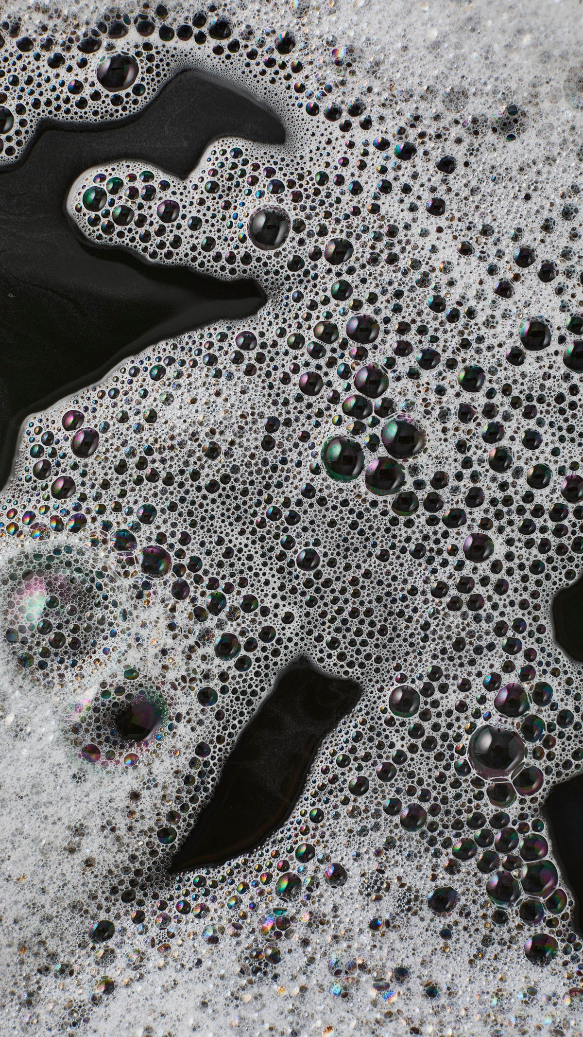 An abstract macro shot of thick white foam floating on dark, near-black bath water. The frothy surface is densely packed with a mixture of tiny white bubbles and larger, translucent bubbles reflecting iridescent colours.