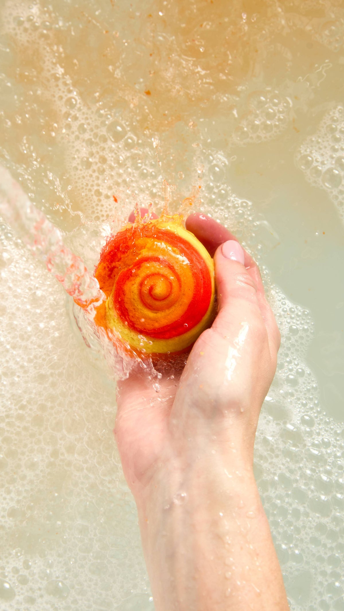 The image is an aerial view of the model's hand reaching in from below and holding the vibrant, orange Brightside mini bubble bar in the hand under running water into the bath water below. Underneath, the water has a layer of thick bubbles.