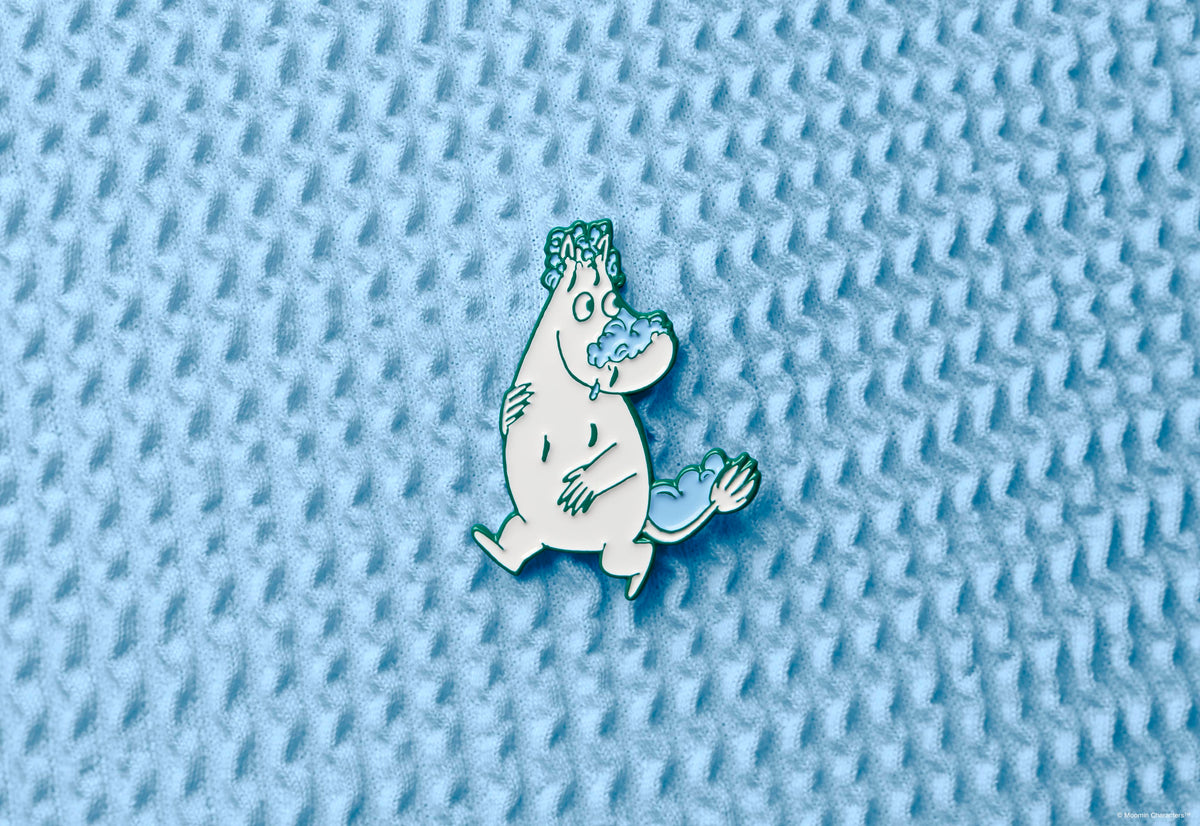 A Moomintroll enamel pin badge, showing the white character covered in blue soap suds on his nose, head, and tail. The pin is displayed in the centre of a textured, light blue waffle-weave fabric background.