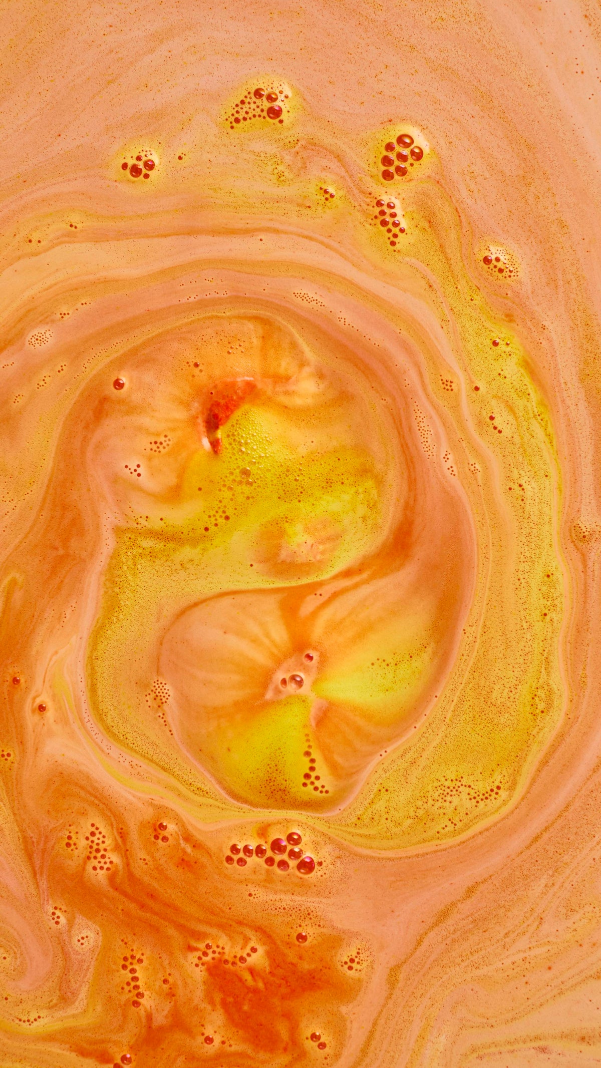 An abstract aerial shot of bath bomb foam swirling in water, creating a large spiral pattern of bright yellow and warm, creamy orange froth. Clusters of fine, orange-tinted soapy bubbles are scattered across the textured surface.